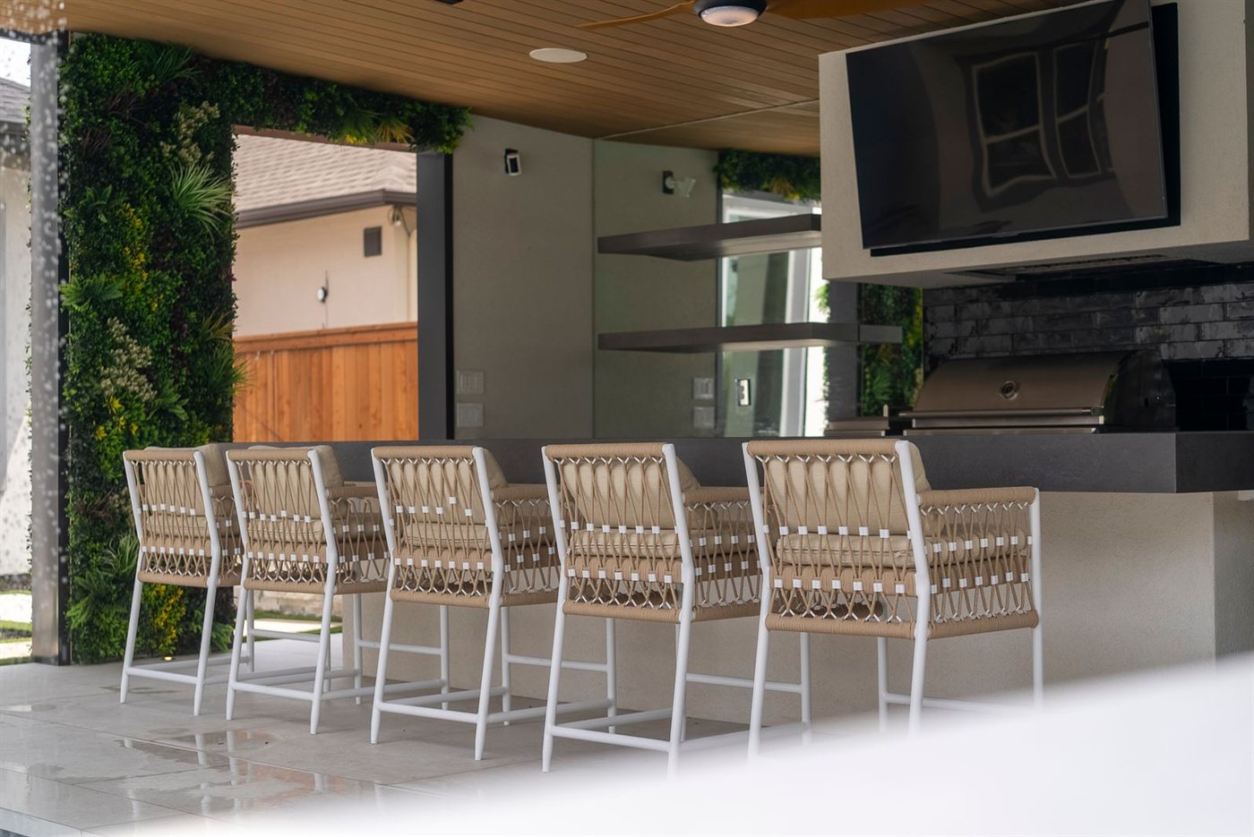 Outdoor bar counter with woven stools beside a grill and wall-mounted TV