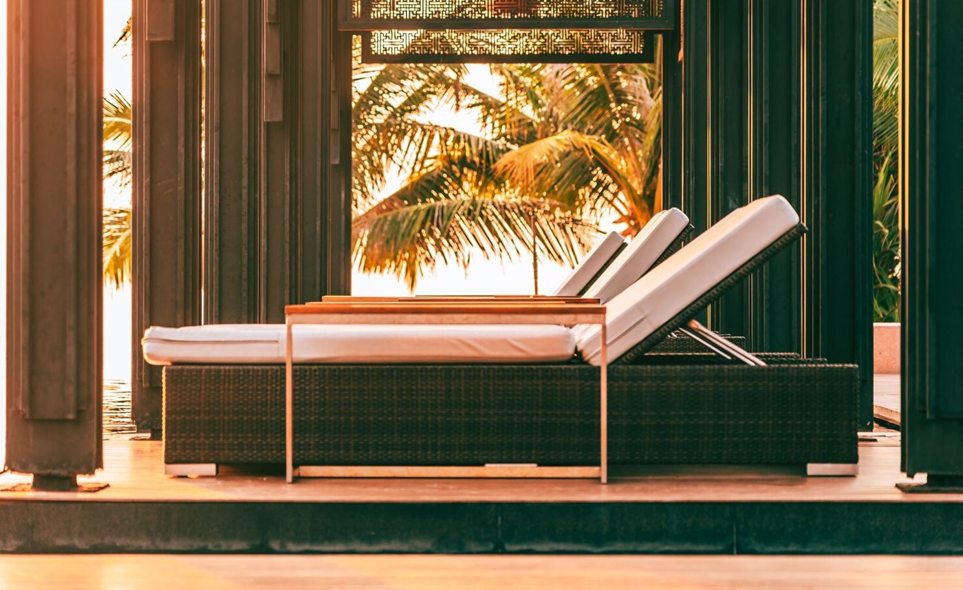 Cushioned lounge chairs under a shaded pergola with palm trees behind