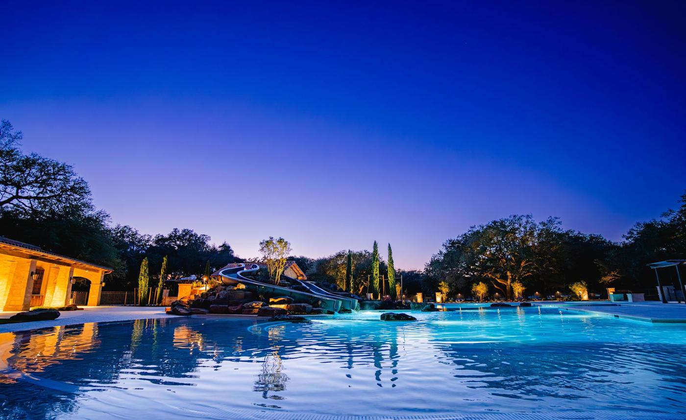 Large resort-style commercial pool with a water slide lit at dusk beneath live oak trees